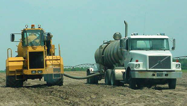 A tanker truck and a spray truck, linked by a hose, in a farm field.