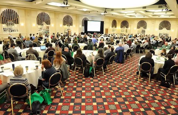 A hotel ballroom room filled with conference attendees watching a presentation. 