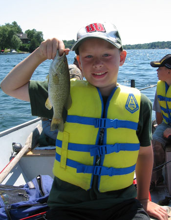 A young boy on a boat wearing a personal floatation device holds up a fish he has caught..