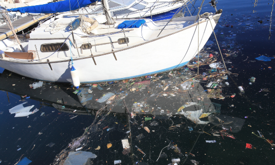 A small white sailboat anchored in water and surrounded by floating trash.