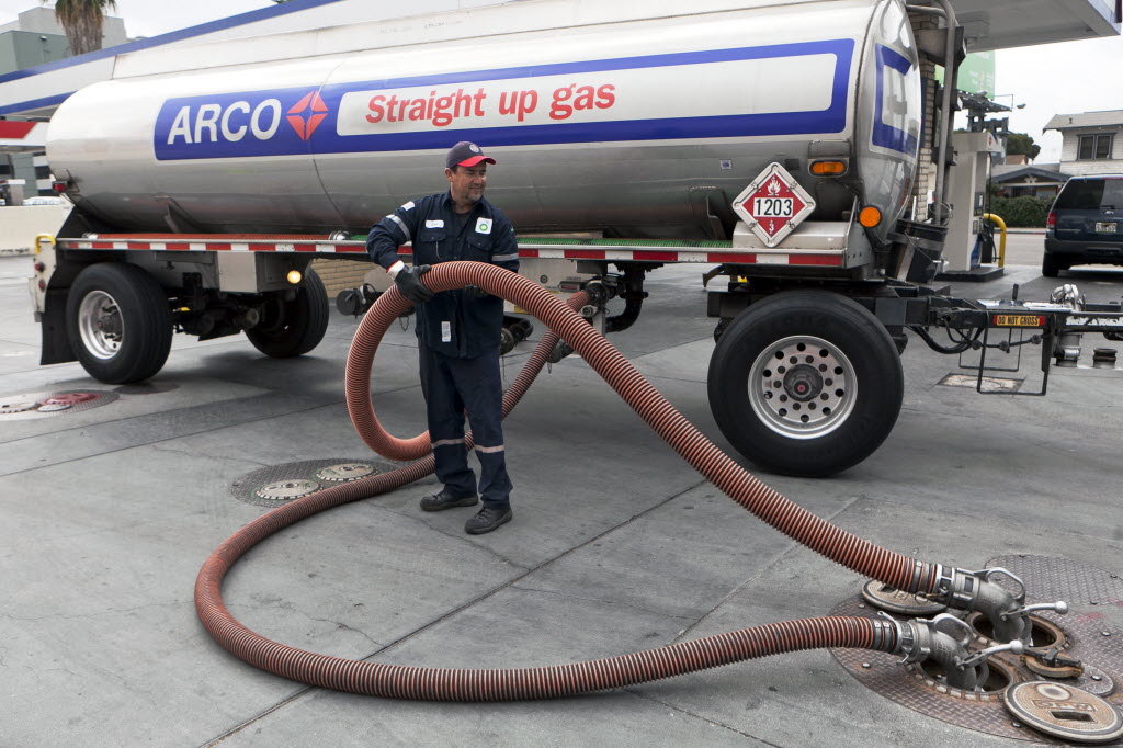A gasoline delivery person holds a hose that leads from a tanker truck to a manhole leading to an underground storage tank.