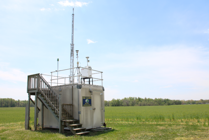 A small block building with a series of sensors and monitors mounted on it sits at the edge of an open field.