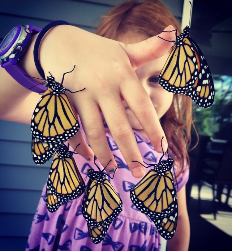 A young girl holds her hand up to the camera. Five butterflies hang from her fingers. 