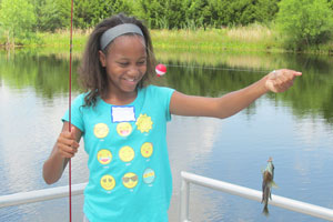 A young girl with fish holds a fishing pole and a fish she has just caught with a pond in the background.