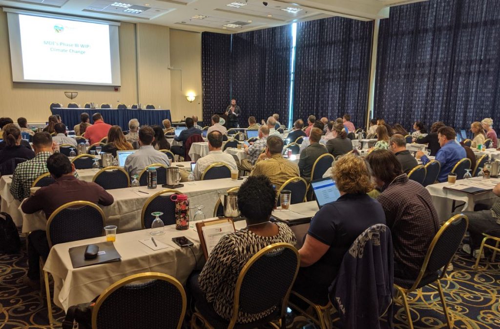 A group of conference attendees, seated at tables, listens to a speaker.