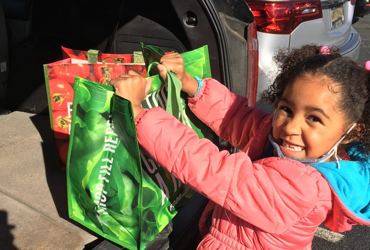 A smiling child loads groceries, packed in reusable shopping bags, into the back of a car.