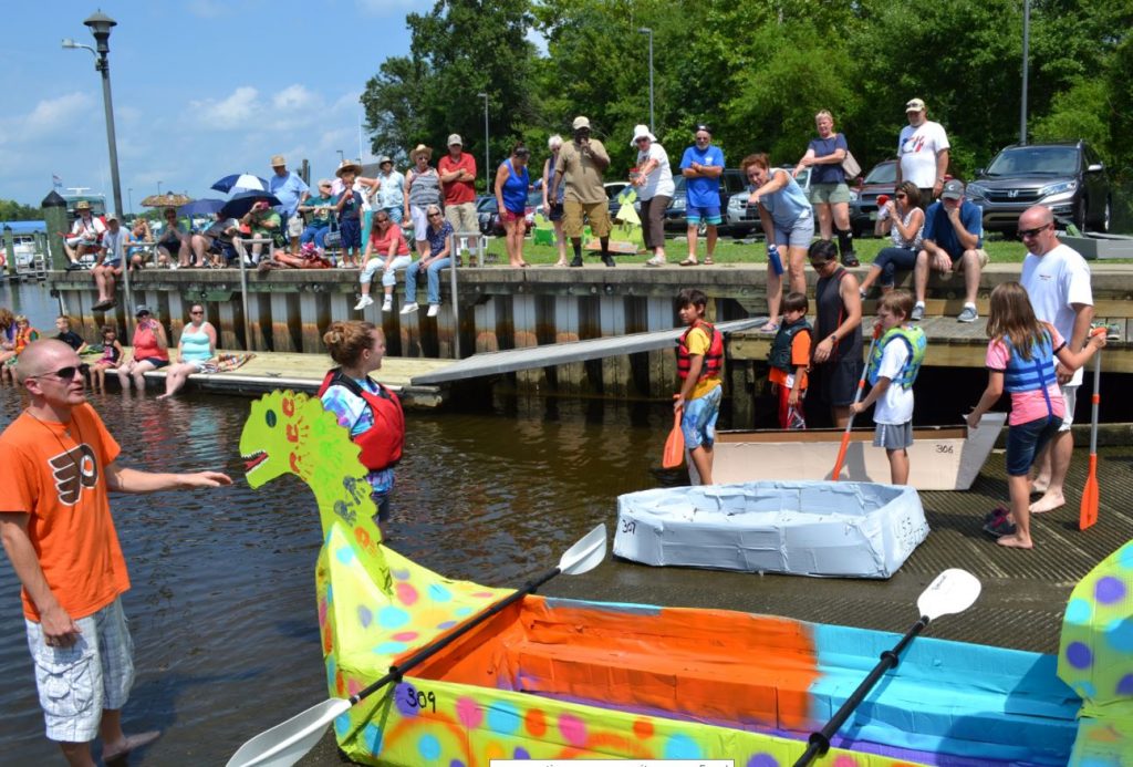 A crowd of people, young and old, at a boat ramp with homemade cardboard boats. 