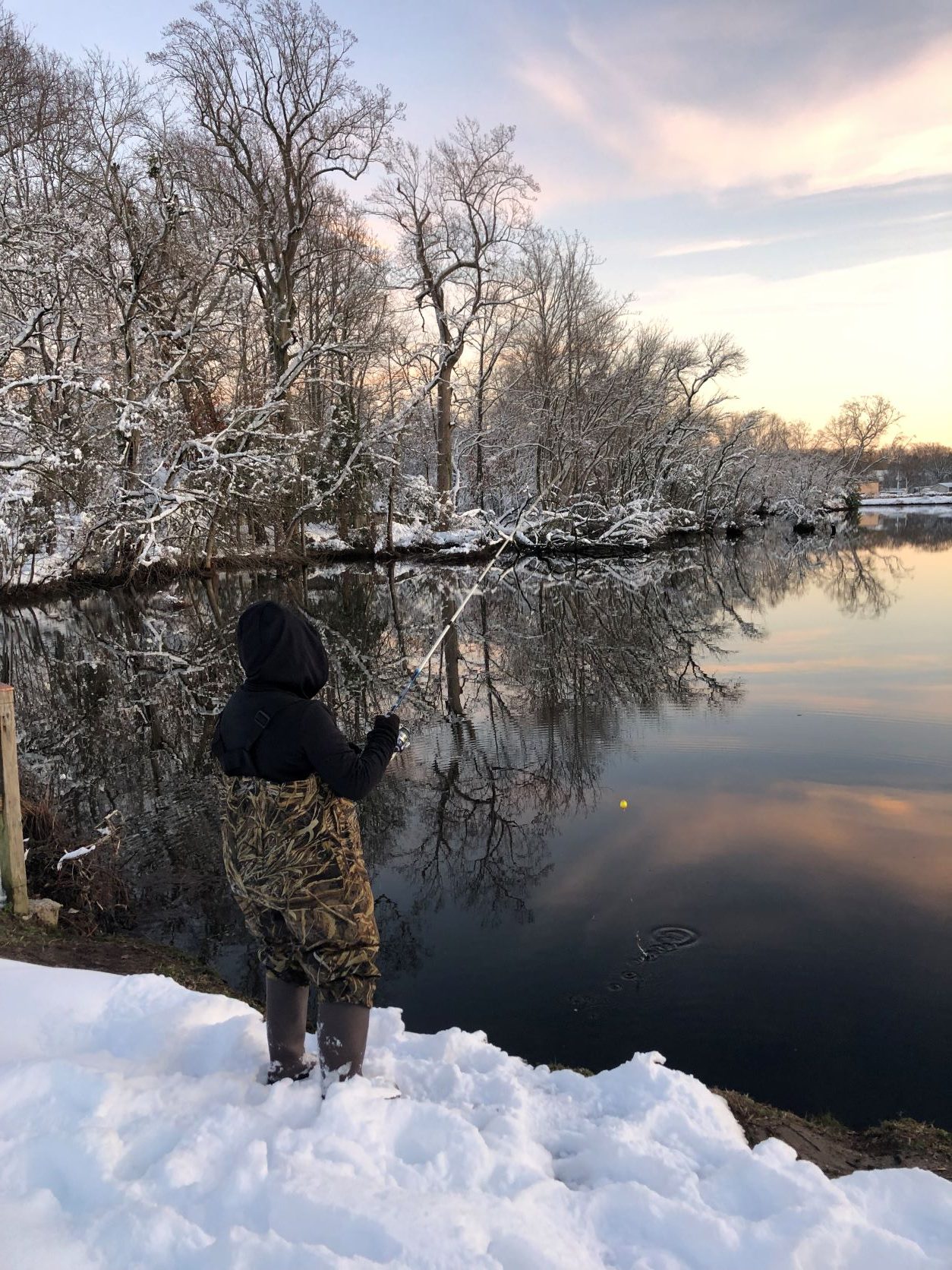 A child dressed in waders fishes from a snow-covered pond shore. 