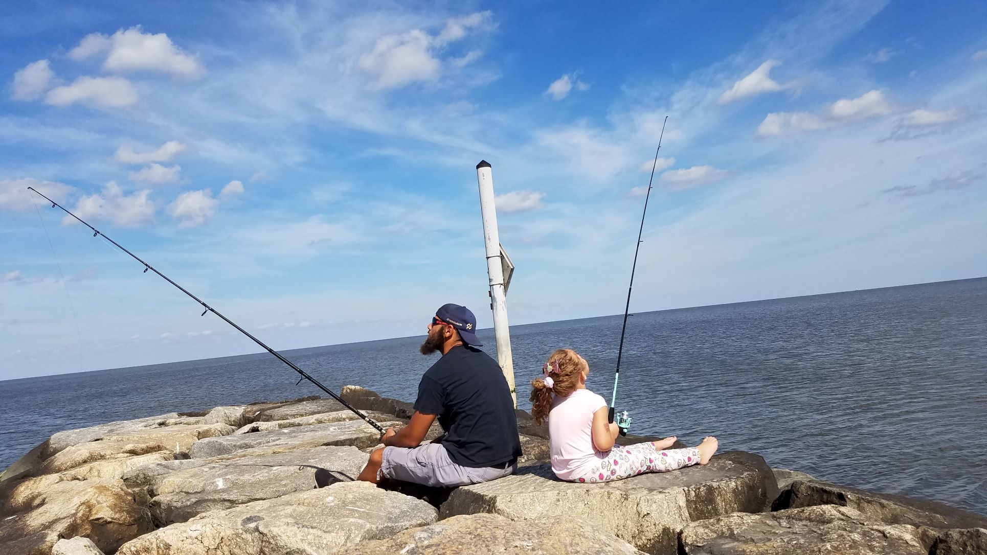 A man and a child sit back-to-back on a stone jetty holding their fishing rods.