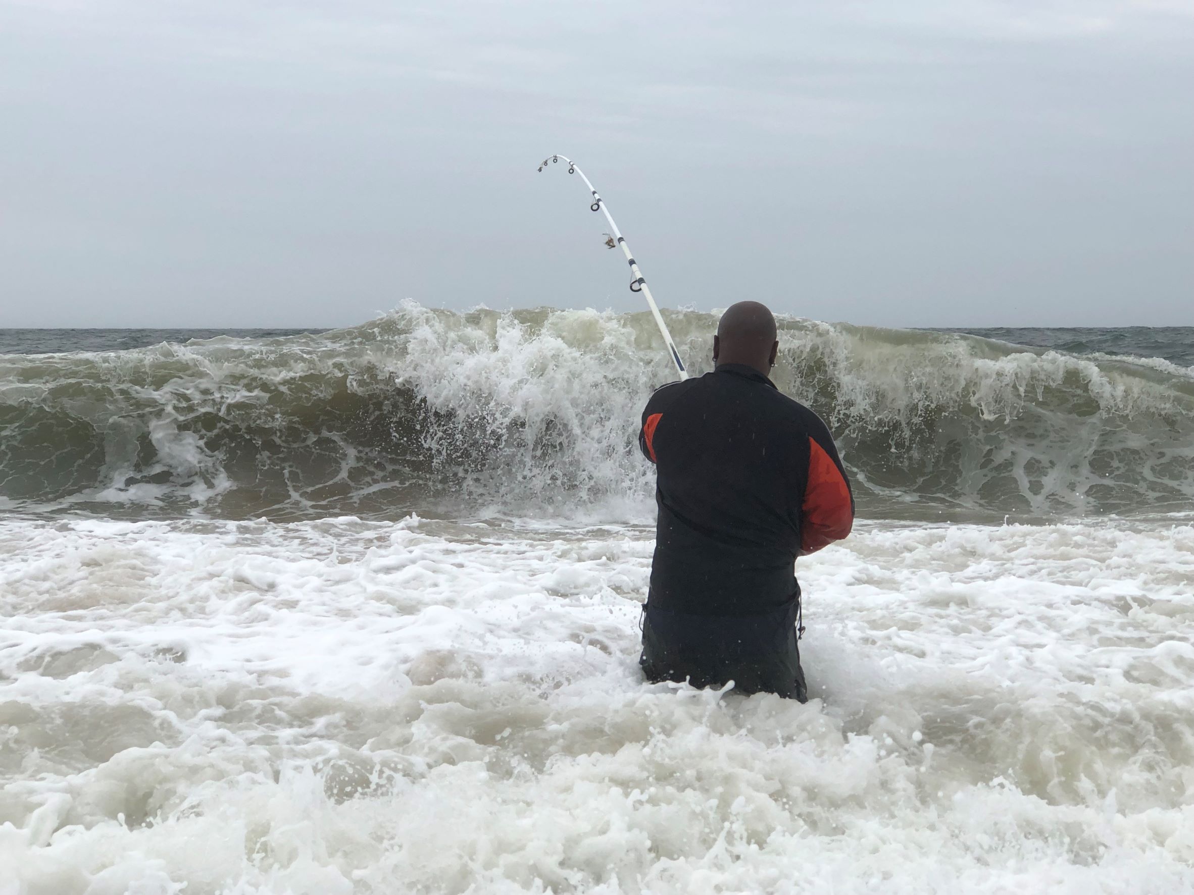 A man stands thigh-deep in ocean water, facing a breaking wave and holding a fishing rod. He appears to be fighting a fish.