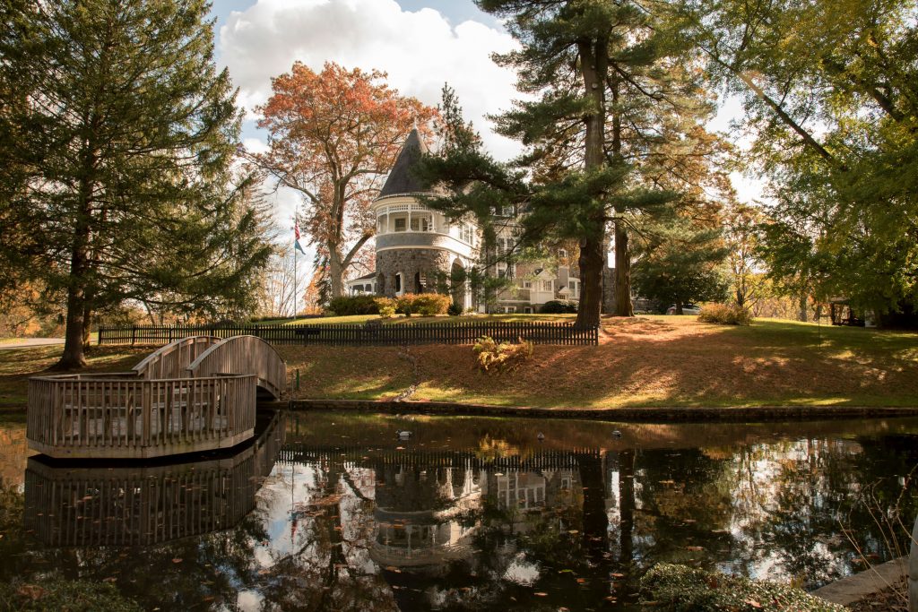 A well-maintained Victorian mansion overlooks a still pond in a wooded setting. 