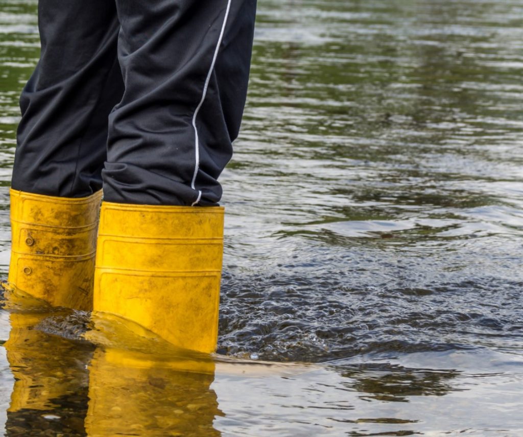 A person's lower legs are seen from behind and to the right, wearing yellow rubber boots that are partially submerged in water.