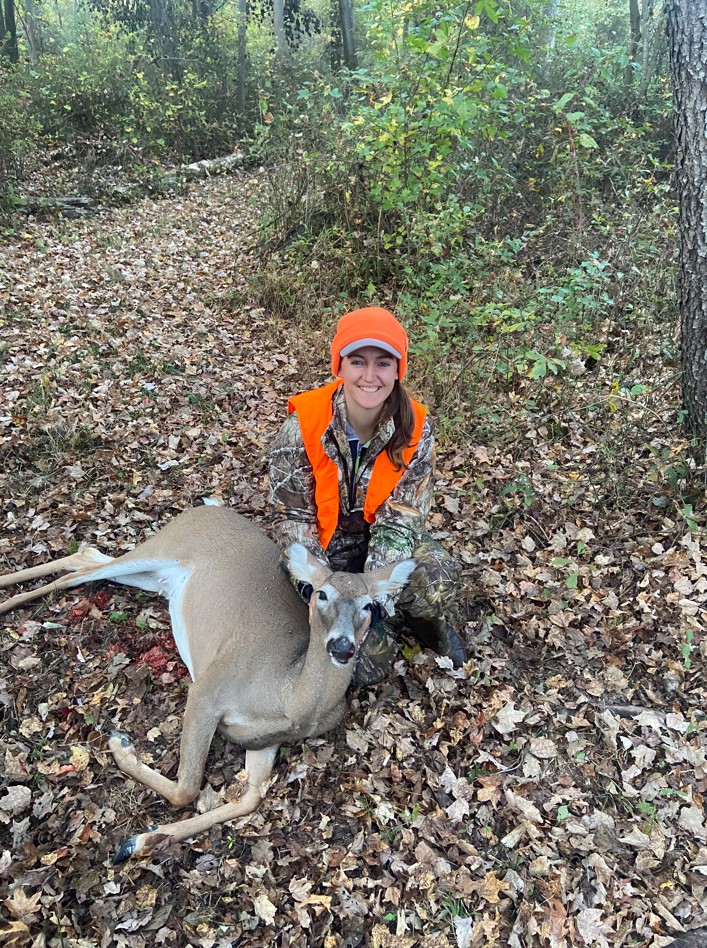 A young woman wearing camouflage clothing, and a vest and hat in hunter orange, poses with a dead doe.