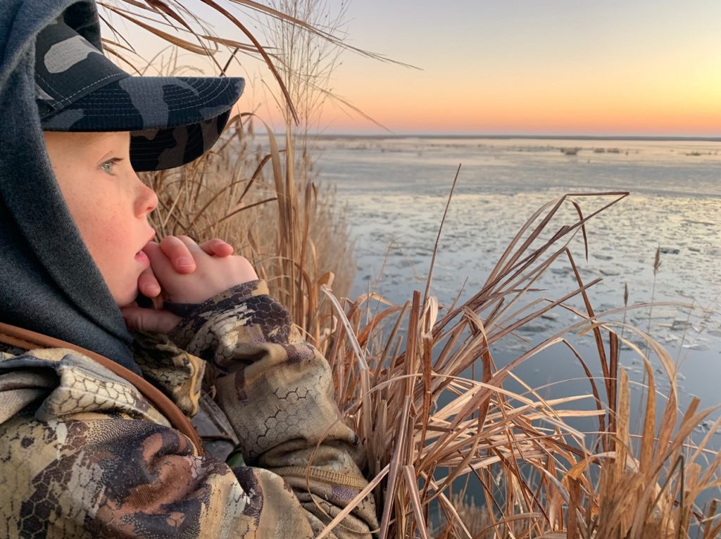 A young boy, bundled up in cold-weather hunting gear, looks out across a winter marsh.