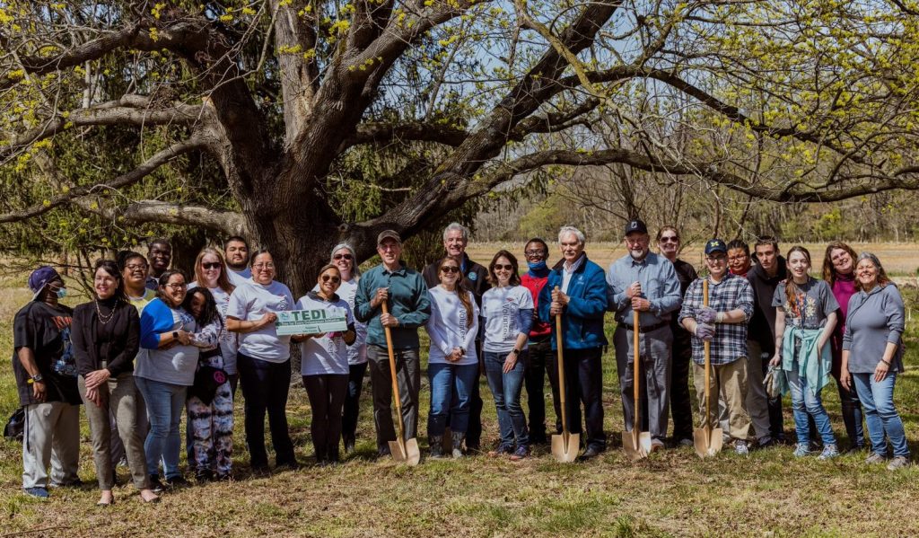 A large group poses with shovels in front of a mature tree.