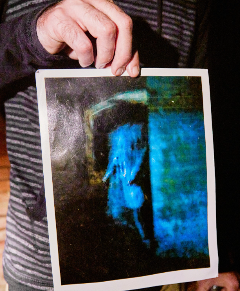 A person holds a photograph that shows a blurry blue figure in a doorway.