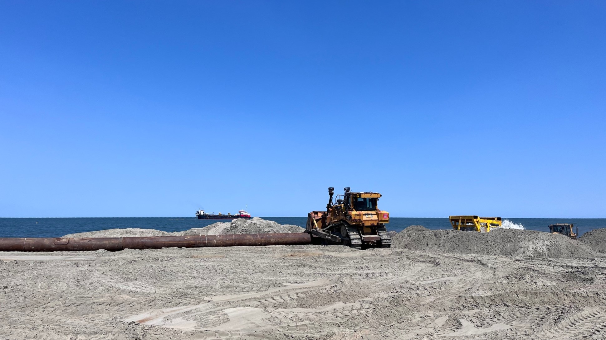 A bulldozer and other heavy equipment move sand that has been pumped onto a beach from a dredge seen floating on the ocean in the distance.