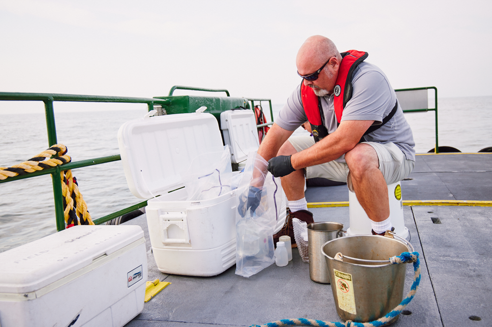 A DNREC environmental control tech wearing a personal flotation device sits on an up-turned bucket on a boat deck and adds water samples to a collection for further analysis.