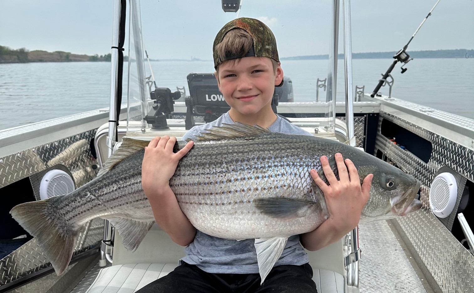 A smiling young angler sits on a boat on the water holding a large fish across his chest.