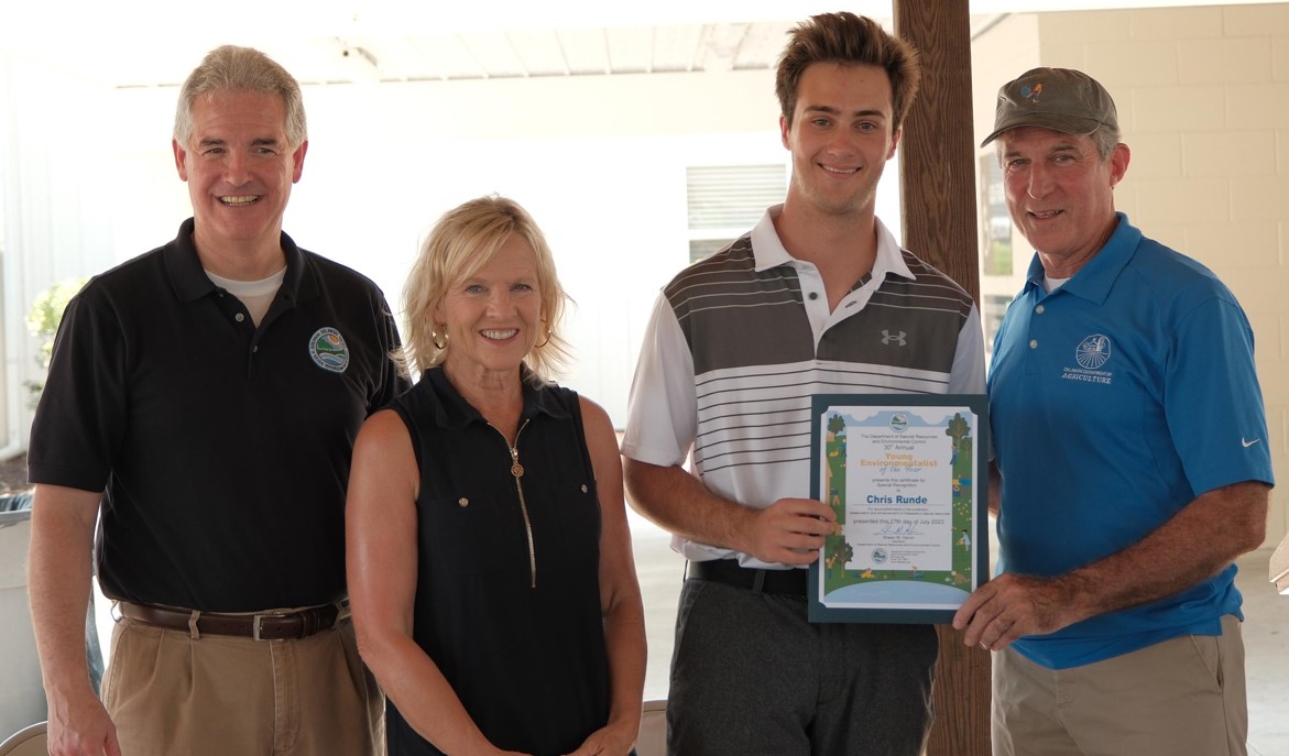 From left to right, a man, a woman, a teenage boy, and a man pose for a photo. The teenager is holding a certificate recognizing his environmental efforts.