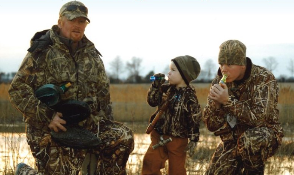 Two men and a small boy are seen in hunting clothes blowing on duck calls in a marshy area.