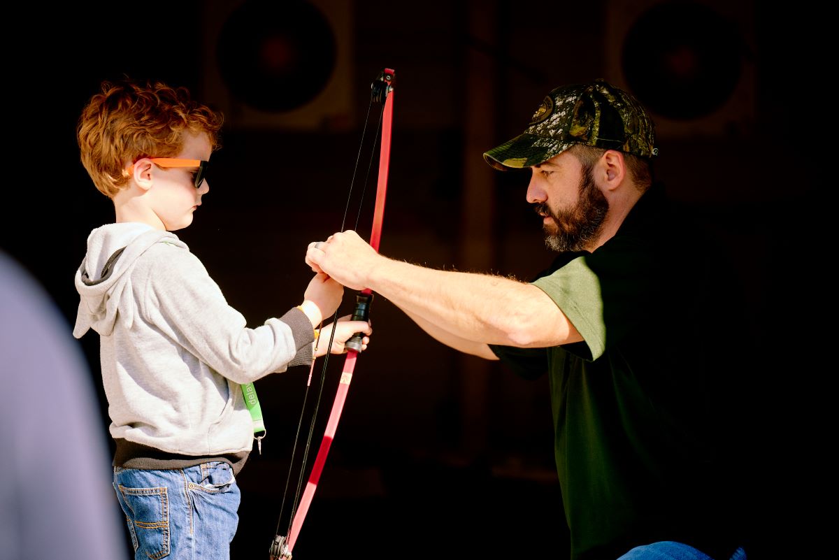 A child learns how to use a bow and arrow, taught by an adult.