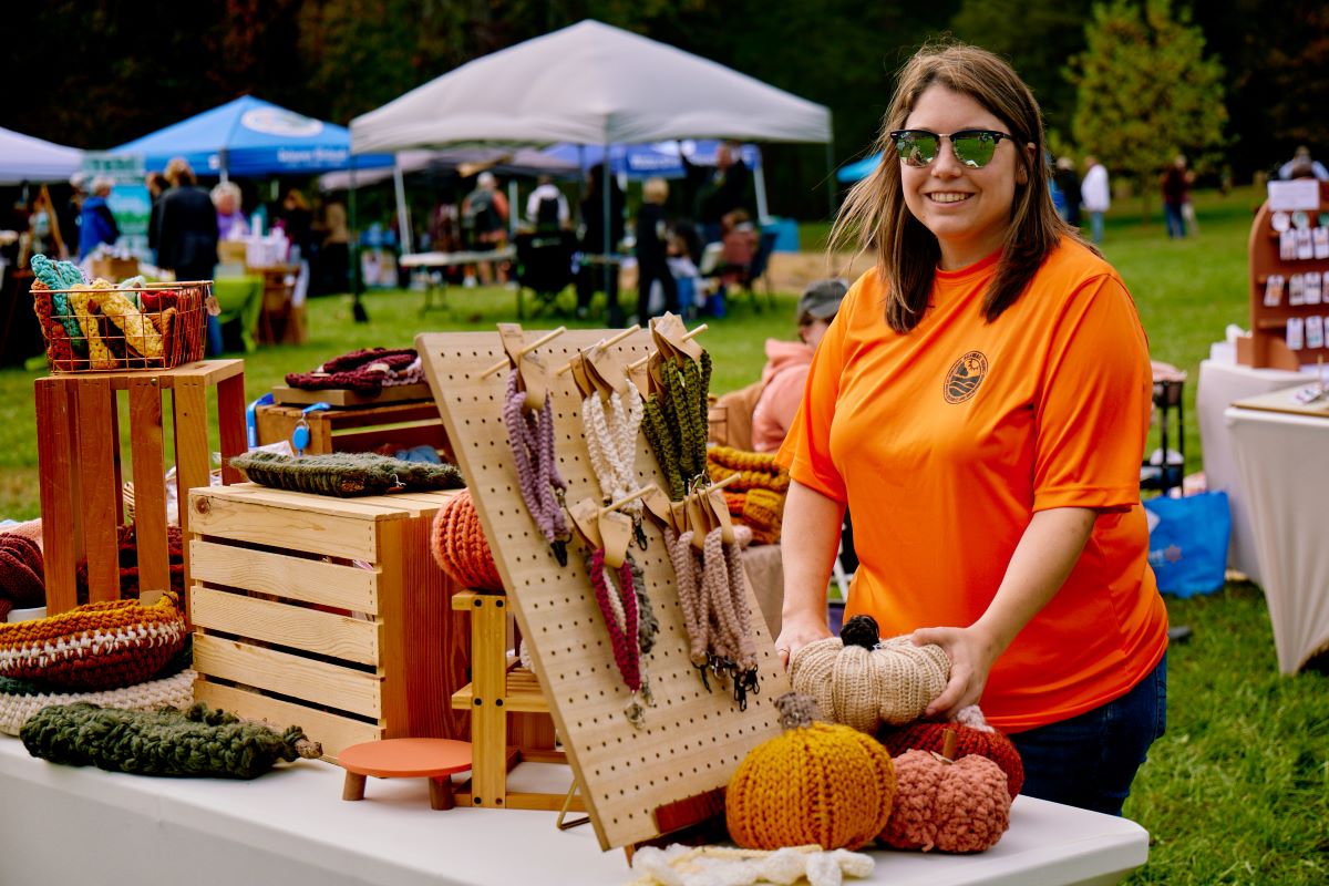 A smiling vendor shows off her crafts at an outdoor festival.