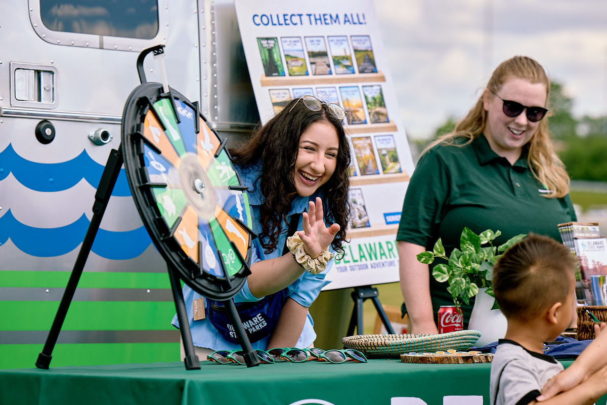 Two adults at a display table at an outdoor event smile as they talk to young children.