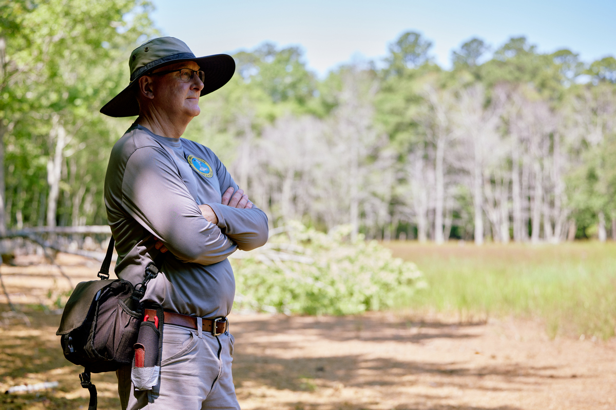 A man dressed for field work in summer weather stands in a field by a line of trees.