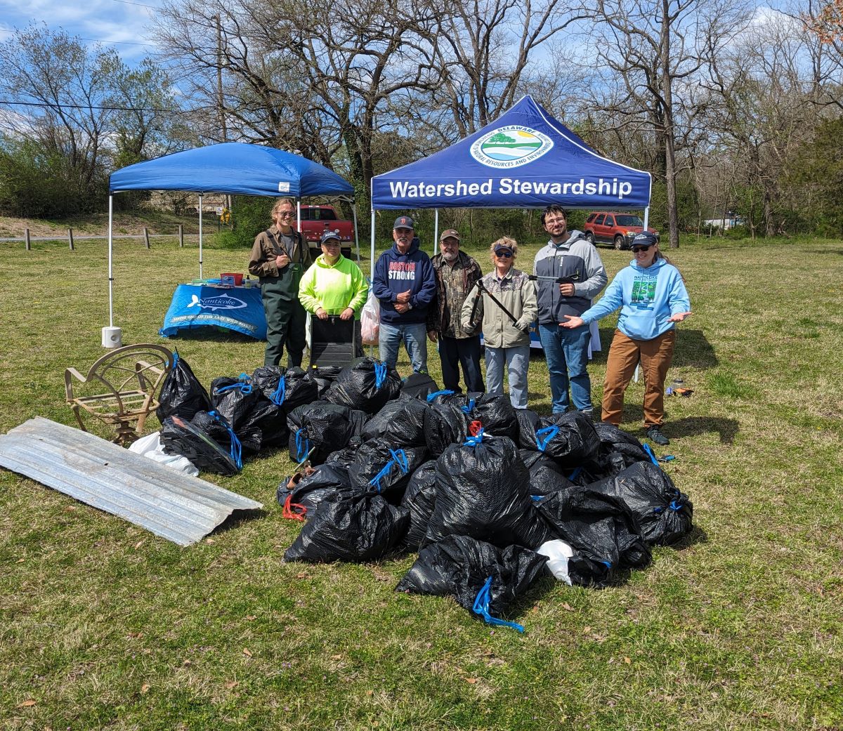 A group of seven volunteers pose behind a large pile of trash bags and debris they have cleared from a pond.