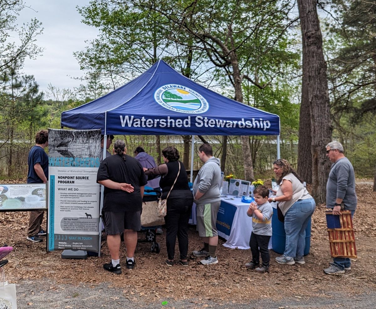 A group of visitors stand next to a blue tent, labelled "Watershed Stewardship" that is set up in front of some trees with a pond in the background.