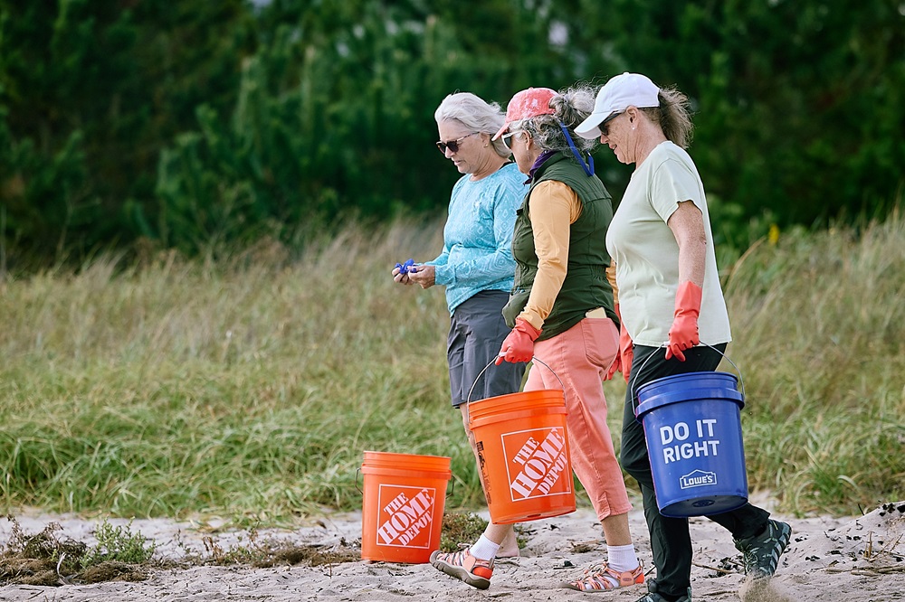 Three women carry colorful buckets from home improvement stores as they walk on a beach.