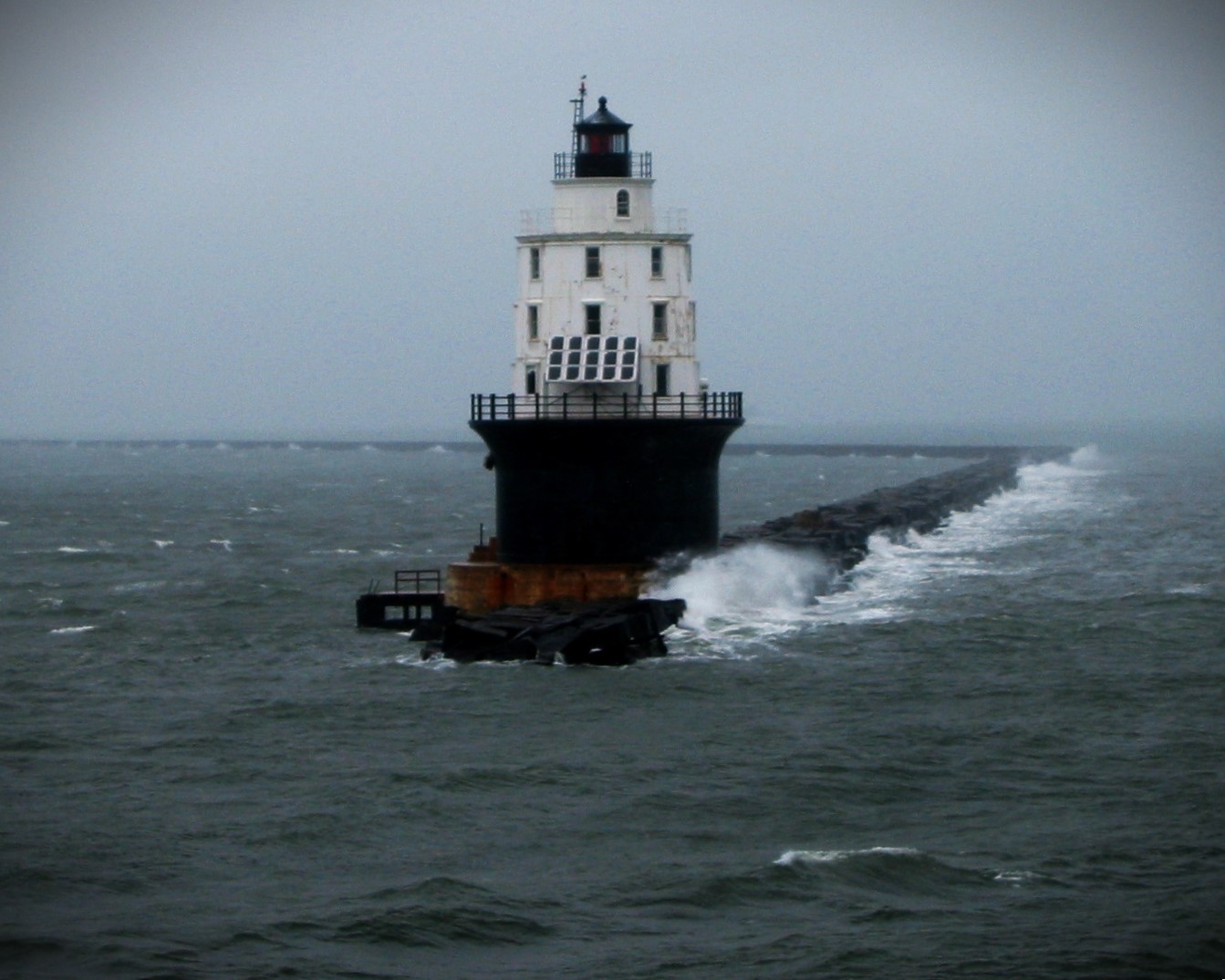 A steel lighthouse is seen perched on the end of a long stone breakwater, with stormy seas breaking on the rocks.