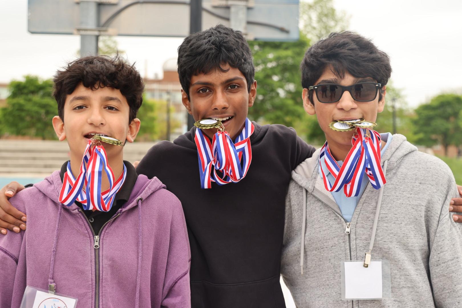 Three students, teenaged boys, pose with their competition medals held in their mouths.