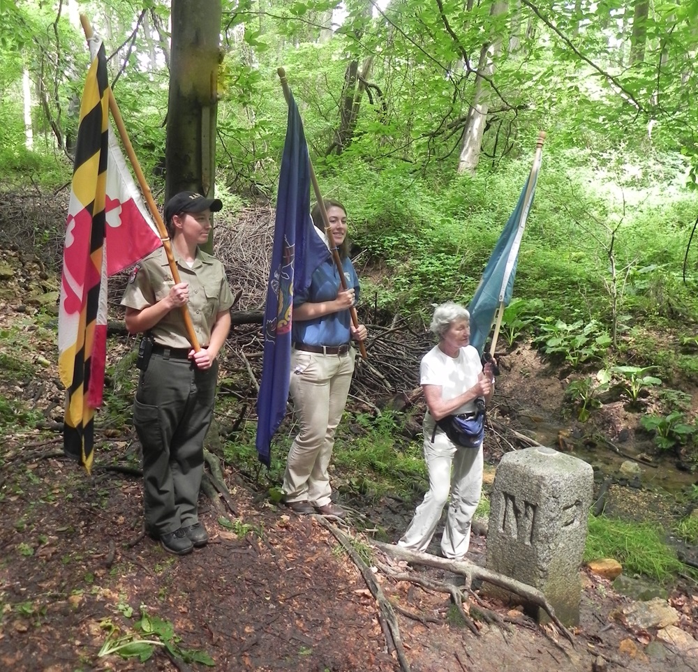 Three people hold state flags alongside a stone marker in a forest.