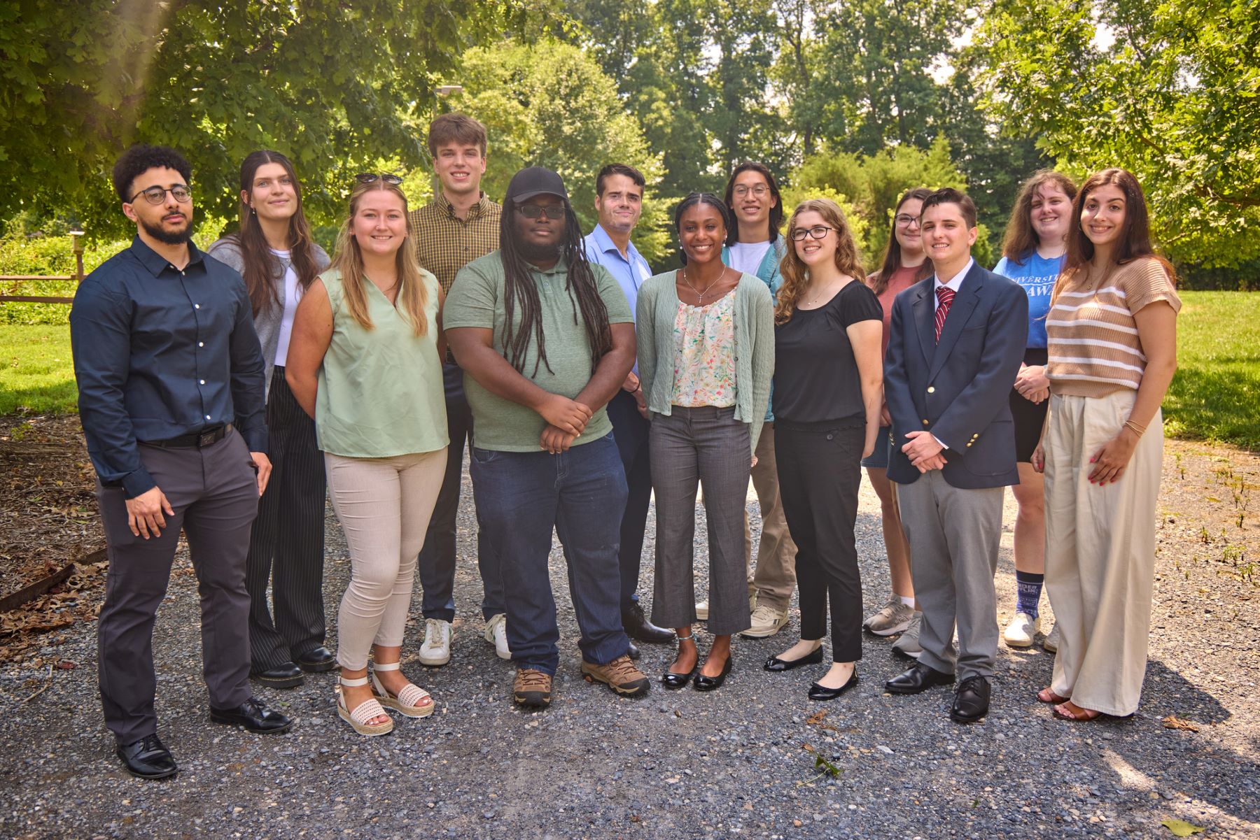 A group of 13 young adults pose outdoors under leafy trees.