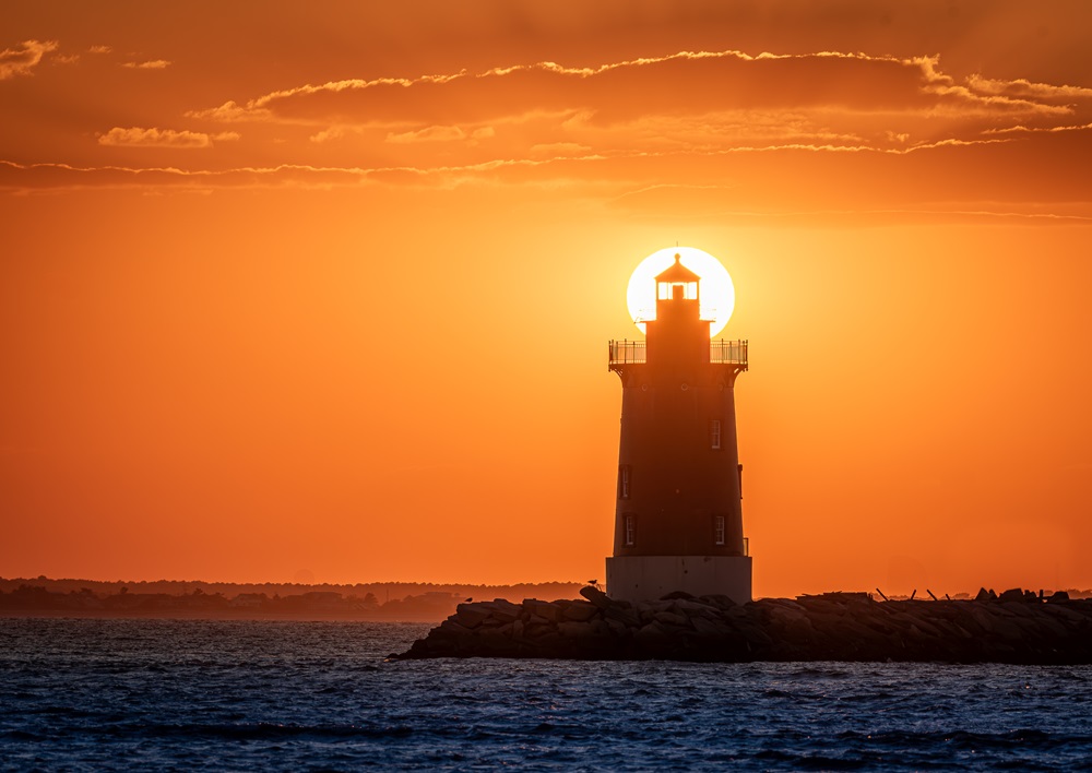 A lighthouse seen at sunset with the sun setting behind it.