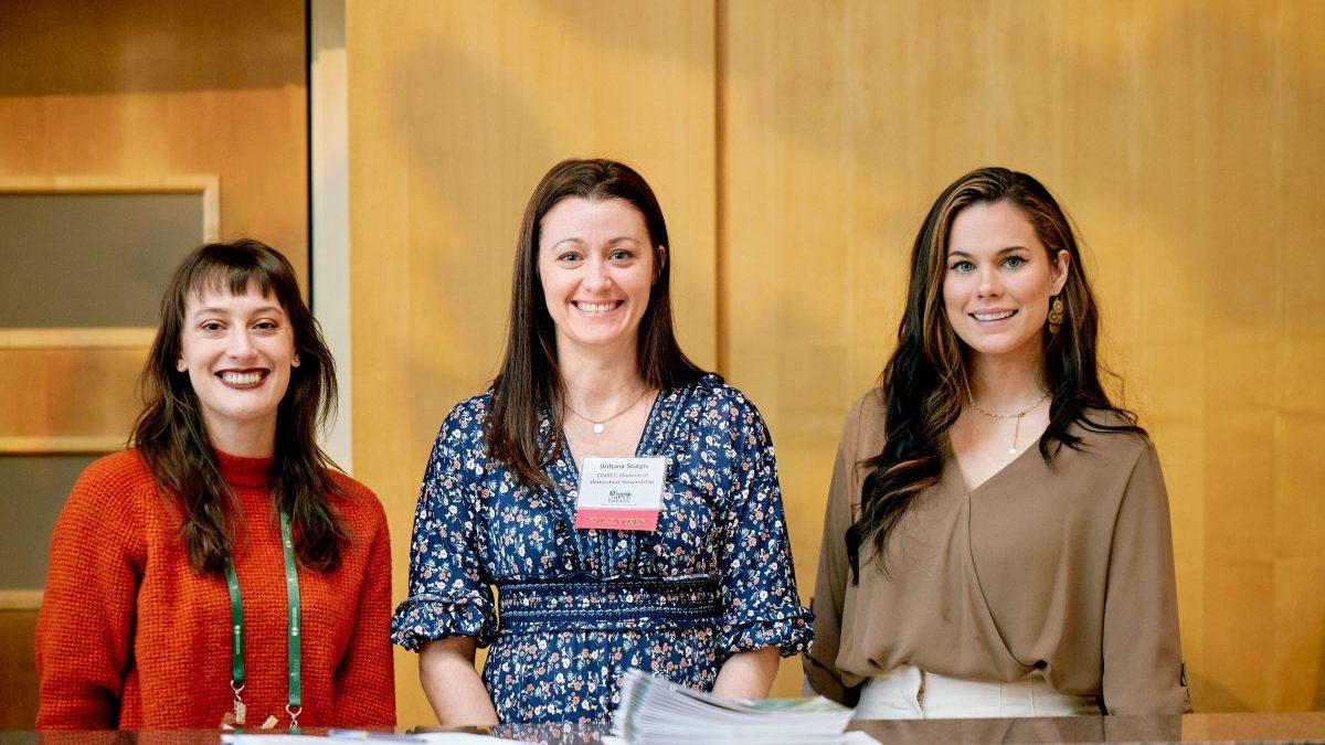 Three smiling women pose in front of a wood panel.