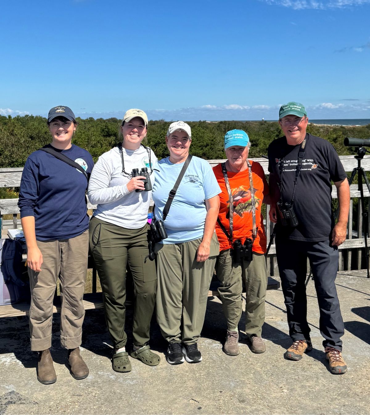 Five adults, some holding binoculars, pose in front of dunes and the ocean.