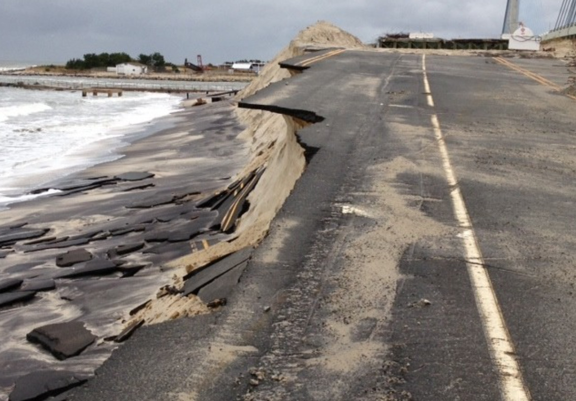 A road along a shoreline crumbles and falls down on the sand at a lower level near the waterline.