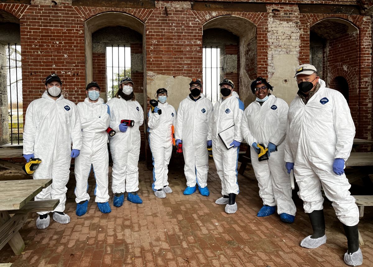 A group of adults wearing white clean-suits and wearing face masks pose in a room that appears to be part of an old brick fort.