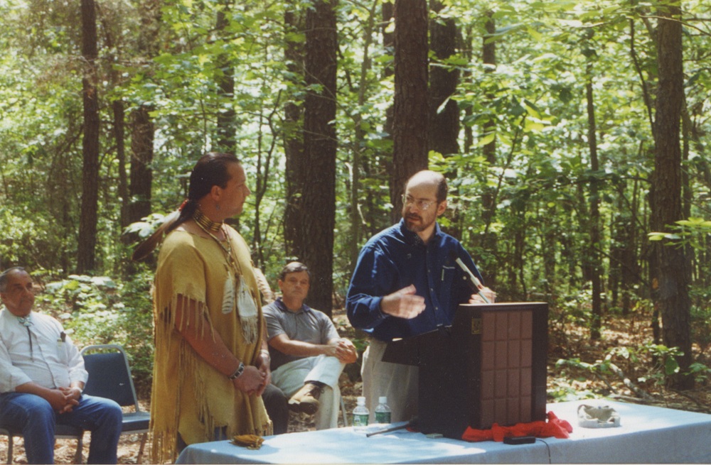 A man stands at a lectern in a forested area, with a man in traditional indigenous garb next to him and several individuals sitting behind him.