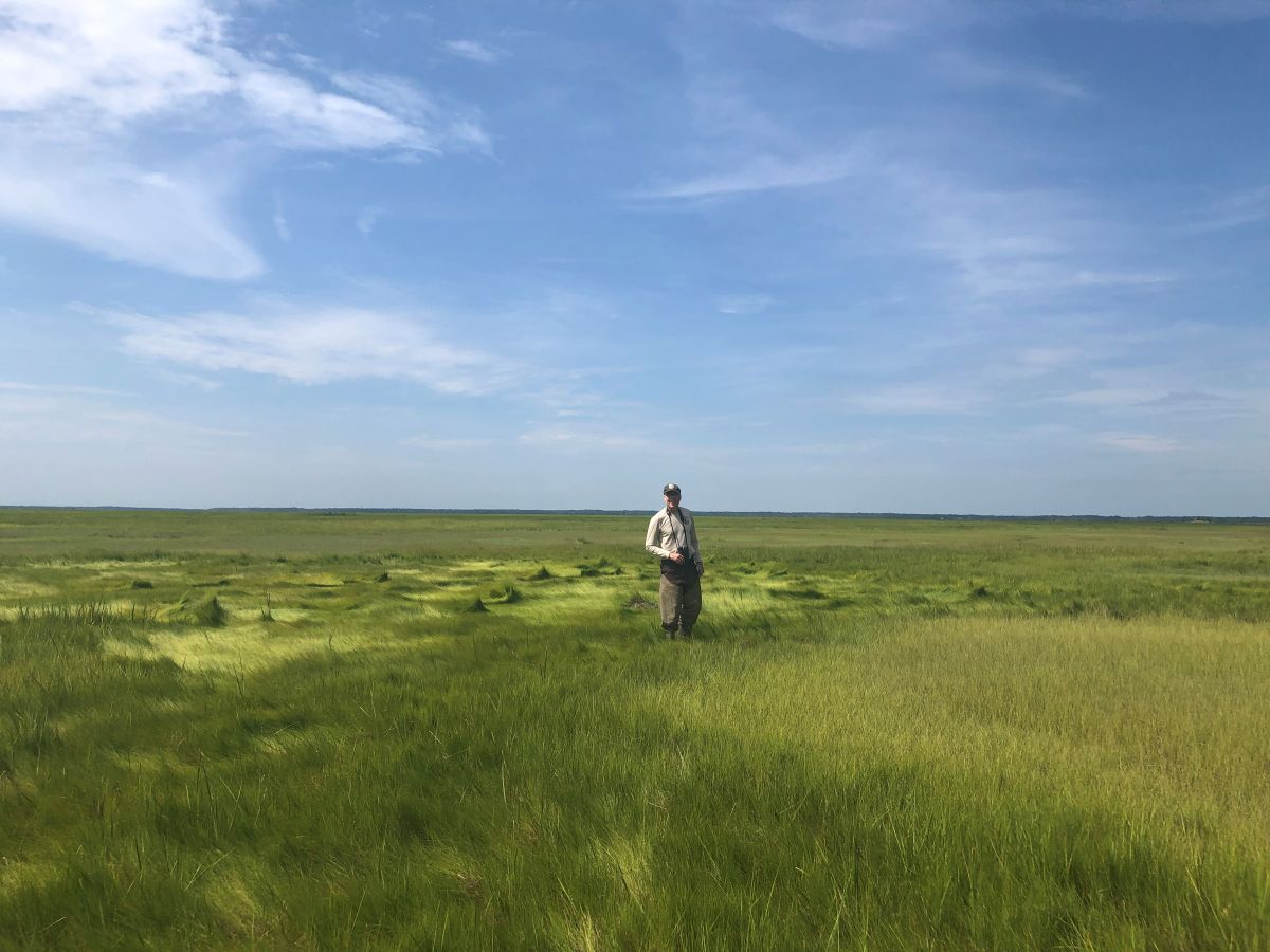 A man stands in a flat, open area of grass holding a pair of binoculars. 