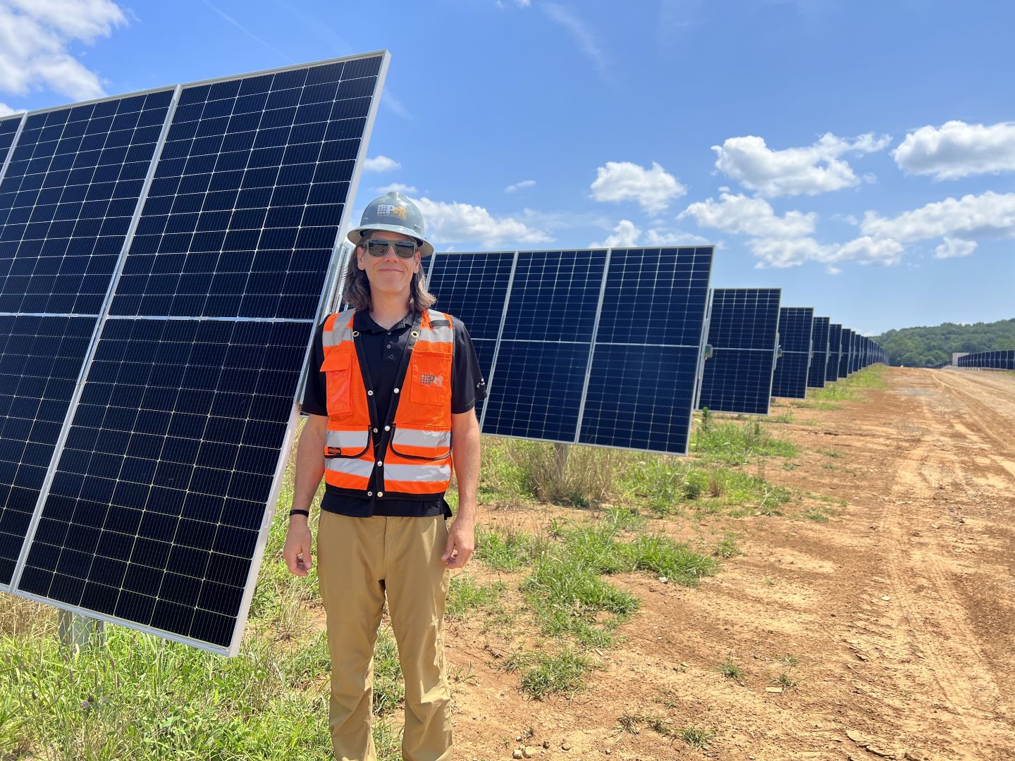 A woman in a hard hat and orange safety vest stands next to solar panels. 
