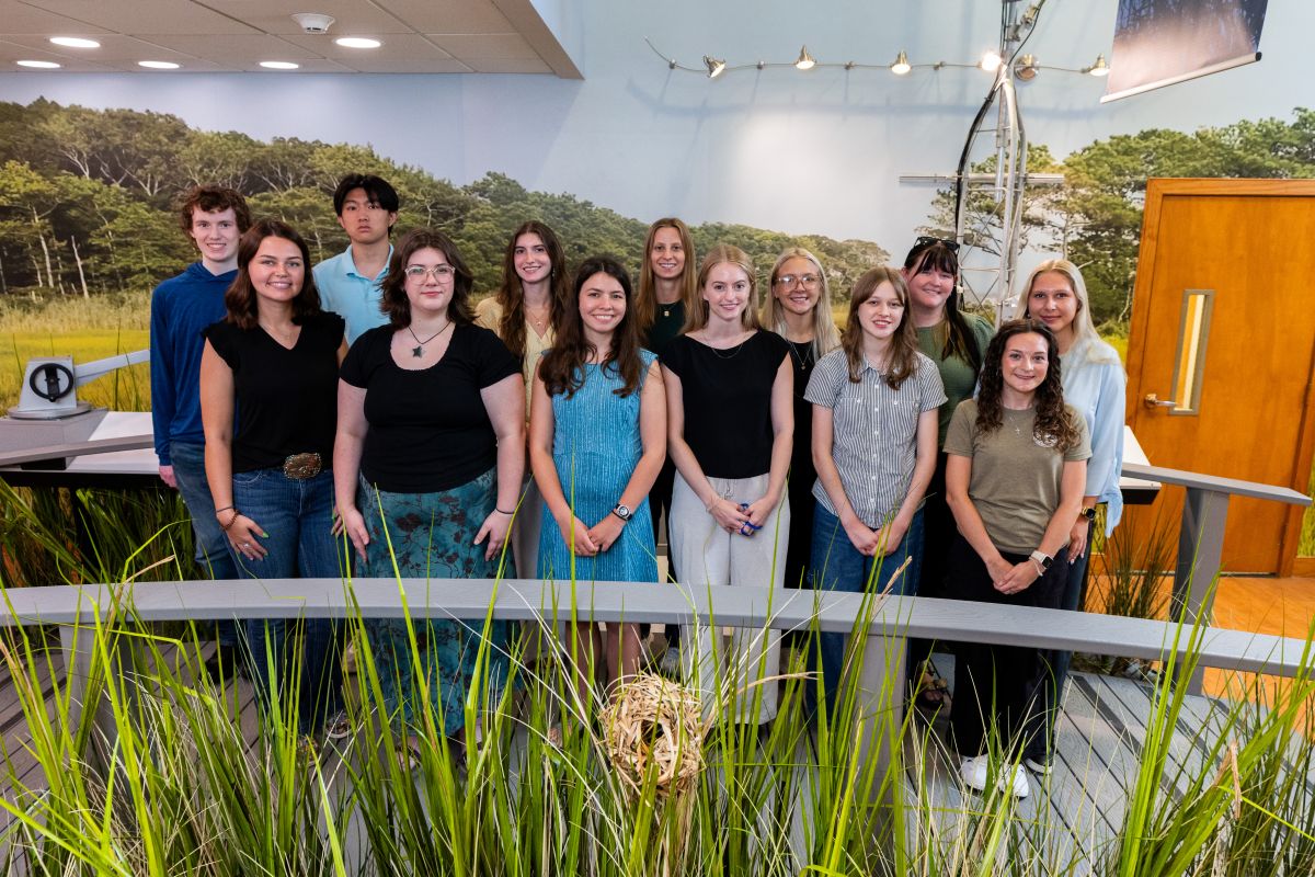 A group of thirteen young people pose together in front of a wall-filling image of a typical Delaware marsh and woodland.