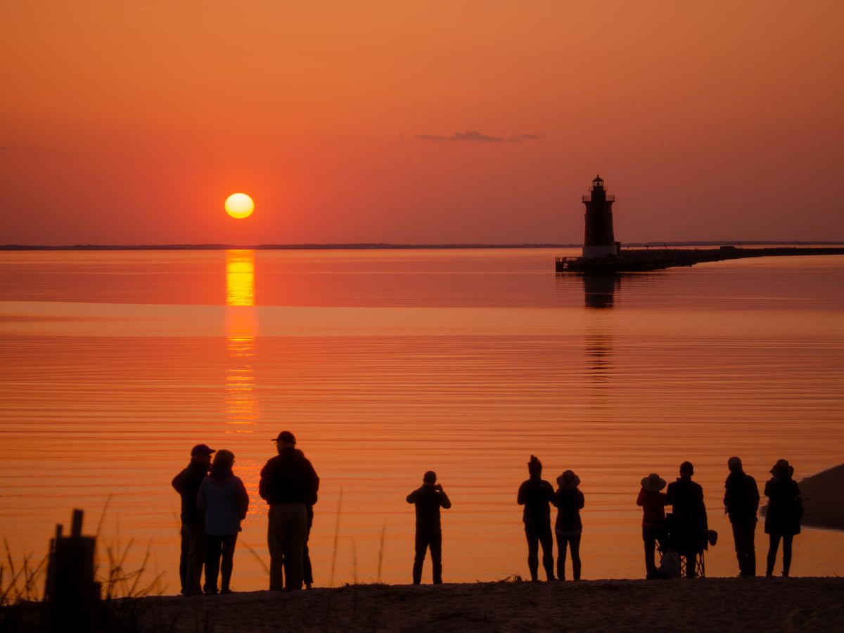 A group of people stand on the shore at sunset looking out towards a lighthouse silhouetted by the setting sun.
