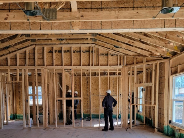 A man in a white hard hat stands in a partially framed-in house.