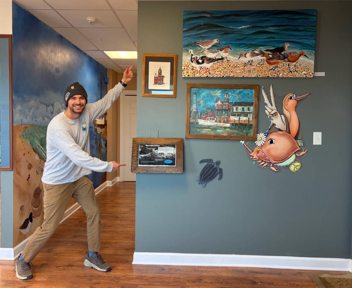 A smiling man points with both hands to a section of wall that has several kinds of art hanging on it.