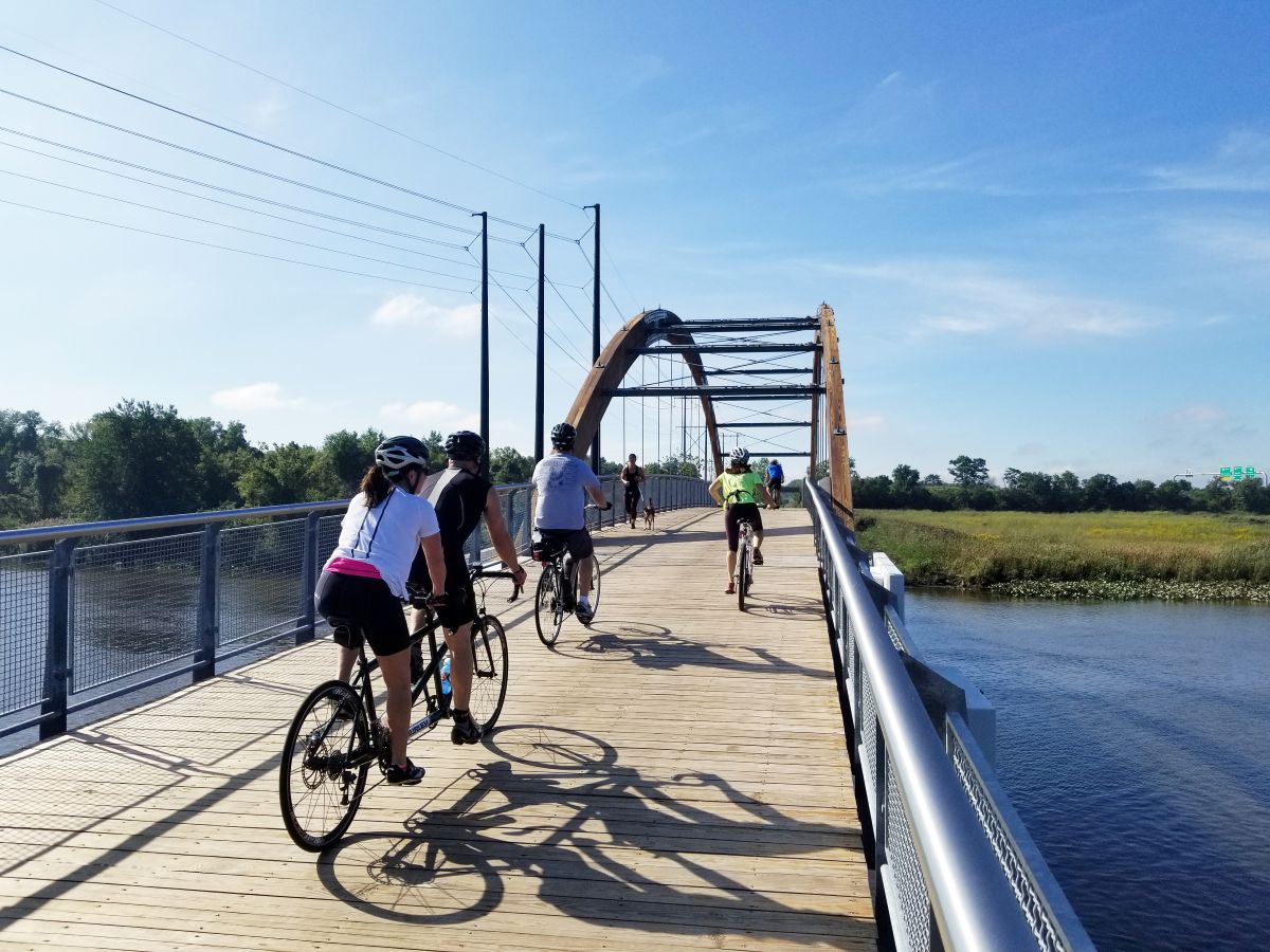 A group of bicyclists cross a stream on a wooden-decked bike bridge.