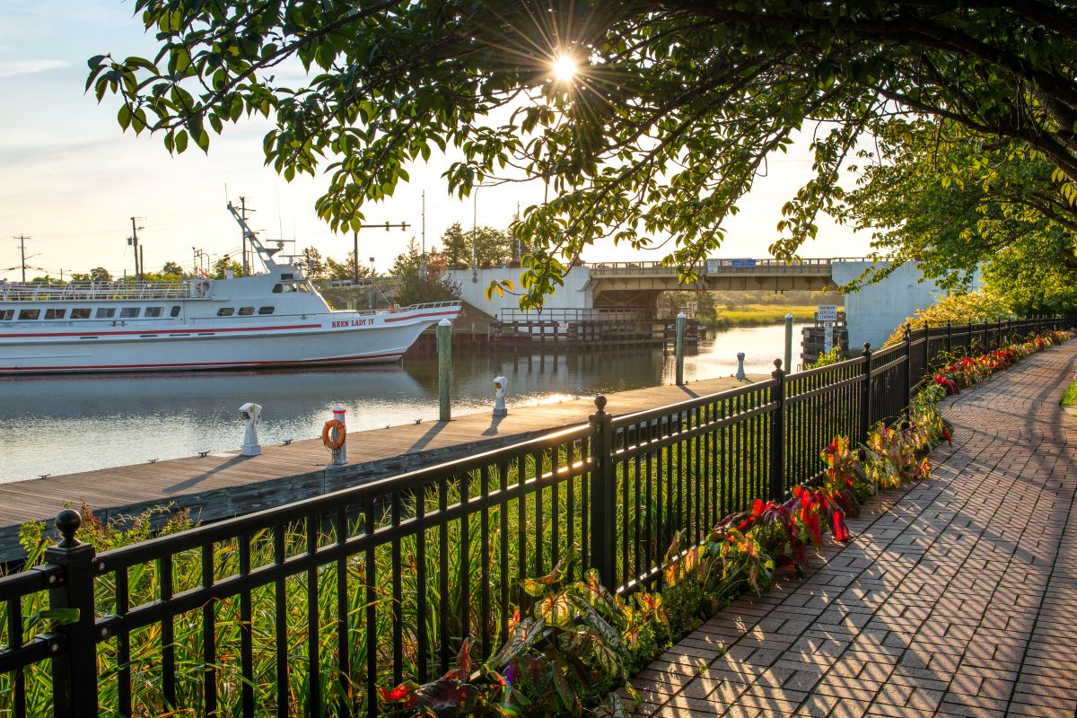 A flower-lined, tree-shaded brick walkway overlooking a small harbor and drawbridge.